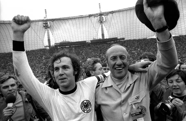 IMAGO Iconic Moments, Fußball Weltmeister Franz Beckenbauer (li.) und Bundestrainer Helmut Schön (re.) jubeln gemeinsam, dahinter ZDF Reporter Dieter Kürten (alle BR Deutschland) Fußball WM Herren Weltmeisterschaft 1974, Nationalmannschaft, Nationalteam, Nationaltrikot, Länderspiel, Finale Gruppe Trainer, Coach, Nationaltrainer, Nationalcoach München Glück, Freude,