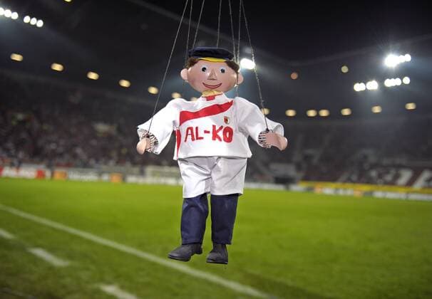 FC Augsburg --- Eine Marionette der Augsburger Puppenkiste, Lukas der Lokomotivführer, mit FCA-Trikot und mit dem Sponsoren-Logo AL-KO auf der Brust hängt im Stadion am Spielfeldrand. o0 kurios Komik Objekte Fan Edelfan Prominente Einzelbild