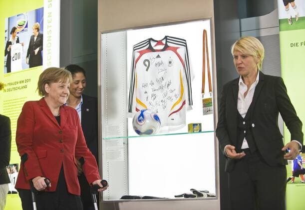 Bundeskanzlerin Angela MERKEL (CDU, li.) sieht sich in der Ausstellung ein Trikot der WM 2007 von Birgit Prinz mit Unterschriften und den Spielball adidas Teamgeist an, daneben Teammanagerin Doris FITSCHEN (re.) und Steffi JONES (WM 2011 OK Präsidentin) --- Eröffnung der Ausstellung FRAUEN SCHREIBEN FUSSBALGESCHICHTE im Bundeskanzleramt Berlin o0 Funktionär Objekte