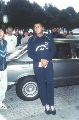 Muhammad Ali (Ex-Boxprofi) 09 84 wot Mann ehemaliger Boxer Box-Profi Profi-Sport Profisport Boxen Weltmeister Boxweltmeister dunkelhäutig dunkelhaarig kurzhaarig leger Sportzeug Sportanzug Jogginganzug blau dunkel dunkelblau Aufschrift Aufdruck Muhammad Ali Name Marke Puma Turnschuhe stehend draussen ganz hoch lächelnd zur Seite schauend sich umdrehend angelehnt an Auto Wagen PKW Unterlagen Zettel haltend Menschen Fans Cassius Clay