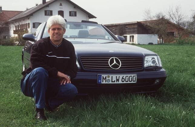 Werner Lorant mit Mercedes 320 SL in auto motor und tv (VOX) 04 00 böw Mann Fußball Trainer Training trainieren TSV 1860 München Fußballverein Verein Spieler Bundesliga Bayern geboren 1948 weißhaarig kurzhaarig leger Sweatshirt schwarz Hose Trainingshose blau vor Auto hockend sich anlehnend rechte Hand auf linke Hand legend ganz quer lächelnd Wiese Haus