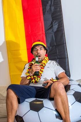 Fußballfan mit Fanartikeln trinkt Bier aus Flasche und schaut Deutschlandspiel auf Fernseher