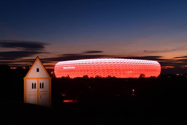 UEFA EURO, EM, Europameisterschaft,Fussball 2020 in Muenchen mit Zuschauer! Archivfoto: ALLIANZ ARENA bei Nacht.Im Vordergrund eine Fakekirche Heilig Kreuz Kirche -Timm Ulrichs, Skulptur aVersunkenes DorfÃ’ im Froetttmaninger Berg. Dynamische Philips LED Beleuchtung sorgt fuer fliesende Uebergaenge in der Lichtanimation. Fussball Arena,Fussballstadion,Stadion,FC Bayern Muenchen,Nachtaufnahme, Abendhimmel,Nachthimmel,rote Beleuchtung,rot beleuchtet,Licht,Stimmung,Abendstimmung,Uebersicht,Totale,Architektur. Â
