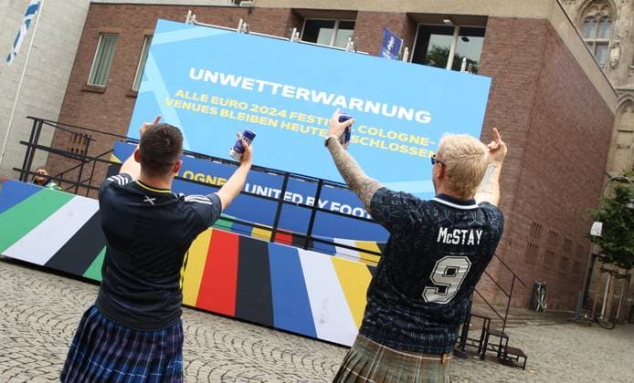 Auf einer digitalen Informationstafel teilt die Stadt Köln mit, dass wegen einer Unwetterwarnung das Fanfest zur Euro 2024 am Dienstag geschlossen bleibt. Diese beiden Fans der schottischen Fußball-Nationalmannschaft lassen sich den Spaß dennoch nicht verderben. Köln Nordrhein-Westfalen