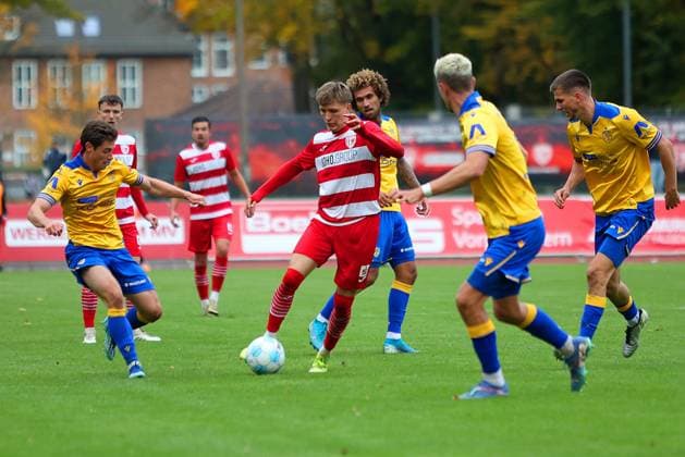 Bild: v.li.: Tristan Wagner 26(Greifswalder FC) vs. Marcel Hoppe 15 und Sören Reddemann 5(FC Carl Zeiss Jena) --- Regionalliga Nordost Greifswalder FC (rot) gegen FC Carl Zeiss Jena (gelb) 25.10.2025 in Greifswald