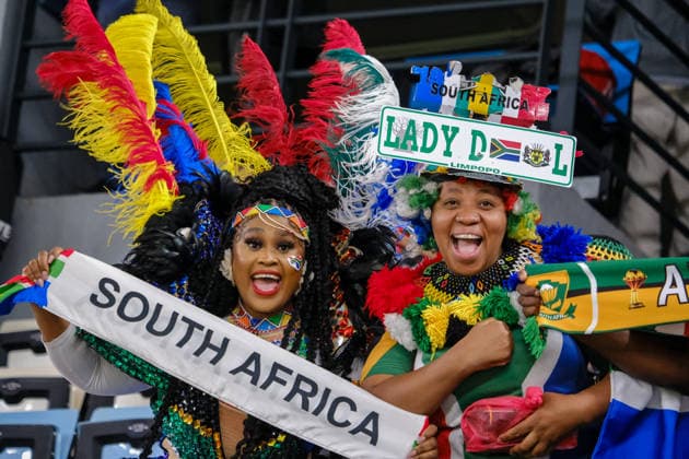 South African fans celebrate in colorful traditional attire at AFCON 2025 RABAT, MOROCCO - JANUARY 04: South African supporters, including one in elaborate feathered traditional dress and another wearing a Lady D Limpopo headpiece, cheer for Bafana Bafana during the AFCON 2025 Round of 16 match between South Africa and Cameroon at Al Barid Stadium on January 04, 2026 in Rabat, Morocco