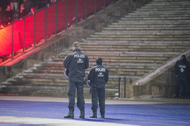 Deutschland: Fussball DFB Pokalspiel 2025 2026, Viertelfinale Hertha BSC Berlin vs SC Freiburg Olympiastadion (Berlin): Zwei Einsatzkräfte der Berliner Polizei am Spielfeldrand mit Schutz- und Einsatzkleidung, Rückenansicht mit Kennzeichnung POLIZEI BE, Absicherung der Stadionbereiche, leere Stehplatztreppen im Hintergrund, rote Pyrotechnikbeleuchtung aus dem Fanblock links, erhöhter Sicherheitsaufwand bei Hochrisikospiel, Flutlichtspiel, Abendspiel, DFB-Pokal, Viertelfinale, K.-o.-Runde, Polizei, Sicherheit, Fußball und Ordnungskräfte, Berlin. *** Germany Soccer DFB Cup match 2025 2026, quarterfinal Hertha BSC Berlin vs SC Freiburg Olympiastadion Berlin Two Berlin police officers on the sidelines with protection and protective clothing, back view with marking POLIZEI BE, protection of the stadium areas, empty standing room stairs in the background, red pyrotechnic lighting from the fan block on the left, increased security during high-risk match, floodlit match, evening match, DFB Cup, quarterfinal, knockout round, police, security, soccer and law enforcement, Berlin Copyright: xJOERANxSTEINSIEKx