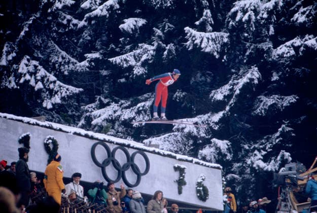 Olympische Winterspiele in Innsbruck 1976 Ein Skispringer in rotem und blauem Anzug mit der Startnummer 50 ist während des Spezialspringens von der 90m-Schanze bei den Olympischen Winterspielen in Innsbruck in der Luft zu sehen. Im Hintergrund sind verschneite Bäume und die olympischen Ringe mit der Jahreszahl 76 zu erkennen. Österreich, 1976 Innsbruck Österreich *** Olympic Winter Games in Innsbruck 1976 a Ski jumper in red and blue Suit with the Starting number 50 is during the Special jumps from the 90m Hill at the Olympic Winter Games in Innsbruck in the Air to see in Background are covered Trees and The Olympic Rings with the Year 76 to recognize Austria, 1976 Innsbruck Austria Copyright: xUnitedxArchivesx xSvenxSimonxSportx SVS_95020000367