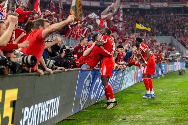 15.04.2026, Fussball UEFA Championsleague 2025 2026: Viertelfinale Rückspiel, FC Bayern München - Real Madrid, in der Allianz-Arena München, Fans stürmen vor Abpfiff den Innenraum und drücken beim Torjubel die Banden mit den Fotografen um, zwei Fotografen werden verletzt, sogar bewusstlos, und müssen danach nötärztlich versorgt werden. Dayot Upamecano (FC Bayern München) umarmt einen Fan