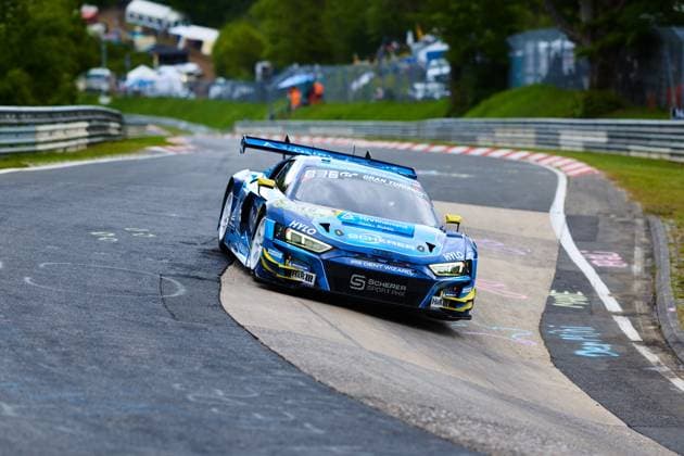 Der Audi R8 LMS GT3 mit der Startnummer 16 von Scherer PHX durchfährt eine enge Kurve auf der Nordschleife des Nürburgrings. Das in Blau und Gelb gehaltene Fahrzeug trägt die Sponsoren HYLO, TÜV Rheinland und H&R und ist mit Michelin-Reifen ausgestattet. Im Hintergrund sind die charakteristischen Leitplanken und das grüne Umfeld der Eifel zu sehen. Alexander Sims & Ben Green & Christopher Haase (Scherer Sport PHX) Audi R8 LMS GT3 16 von Scherer Sport PHX auf der Nordschleife