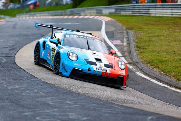 Der Porsche 911 GT3 mit der Startnummer 925 von Huber Motorsport durchfährt eine enge Kurve auf der Nordschleife des Nürburgrings. Das Fahrzeug Christopher Allen & Hans Wehrmann & Jaden Lander & Jake Walker & Jon Miller & Jonathan Miller & Robert Mau (Huber Motorsport) präsentiert sich in einem auffälligen hellblauen und orangeroten Farbschema mit Sponsoren wie H&R und Gran Turismo. Der Bolide liegt tief in der Kurve und zeigt die charakteristische Fahrwerksarbeit des Porsche GT3 auf dem anspruchsvollen Kurs. Porsche 911 GT3 925 von Huber Motorsport auf der Nordschleife