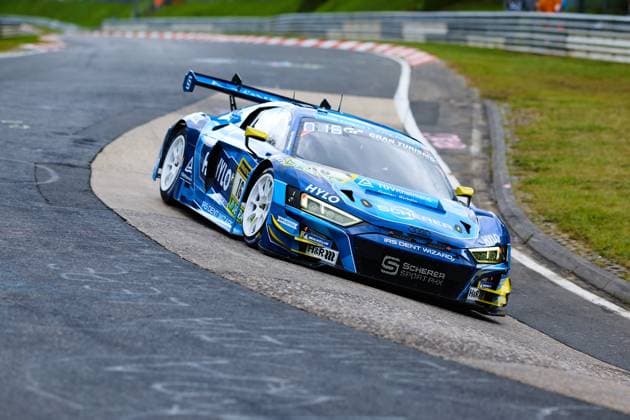 Der Audi R8 LMS GT3 mit der Startnummer 16 von Scherer PHX durchfährt eine Kurve auf der Nordschleife des Nürburgrings. Das in Blau gehaltene Fahrzeug Alexander Sims & Ben Green & Christopher Haase (Scherer Sport PHX) trägt die Sponsoren HYLO, IRS Dent Wizard und TÜV Rheinland prominent auf der Karosserie. Die Michelin-bereiften weißen Felgen und der markante Heckflügel unterstreichen den GT3-Charakter des Audi. Audi R8 LMS GT3 16 von Scherer Sport PHX in der Kurve