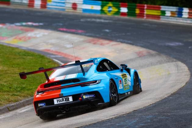 Der Porsche 911 GT3 mit der Startnummer 925 von Huber Motorsport durchfährt eine enge Kurve auf der Nordschleife am Nürburgring. Das Fahrzeug Christopher Allen & Hans Wehrmann & Jaden Lander & Jake Walker & Jon Miller & Jonathan Miller & Robert Mau (Huber Motorsport) ist in einem auffälligen Blau-Rot-Weiß-Design gehalten und trägt Sponsoren wie H&R und Michelin. Im Hintergrund sind die charakteristischen bunten Fahnen und Graffiti-Bemalungen der Nordschleife zu sehen. Porsche 911 GT3 925 von Huber Motorsport auf der Nordschleife