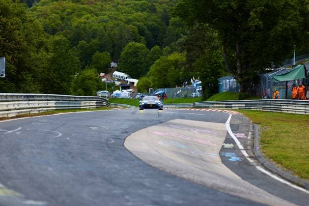 Ein blauer Mercedes-AMG GT3 mit der Startnummer 99 durchfährt eine charakteristische Kurve auf der Nordschleife des Nürburgrings. Im Hintergrund sind die typischen grünen Hügel der Eifel sowie Streckenposten in orangefarbener Ausrüstung zu sehen. Die rot-weißen Curbs und die geschwungene Streckenführung unterstreichen den besonderen Charakter dieser legendären Rennstrecke. Mercedes-AMG GT3 99 auf der Nordschleife am Nürburgring
