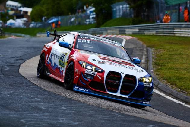 Der BMW M4 mit der Startnummer 145 des Teams Riller & Schnauck powered by Cerny Motorsport durchfährt eine enge Kurve auf der Nordschleife des Nürburgrings. Das Fahrzeug Aris Balanian & Jeroen Bleekemolen & Joshua Bednarski & Peter Cate & Tom Schütze (Riller & Schnauck powered by Cerny Motorsport) präsentiert sich in einem auffälligen rot-weiß-blauen Design mit Sponsoren wie H&R, Michelin und Gran Turismo auf der Karosserie. Im Hintergrund sind Streckenposten und die charakteristischen Leitplanken der Nordschleife zu erkennen. BMW M4 145 von Riller & Schnauck powered by Cerny Motorsport am Nürburgring