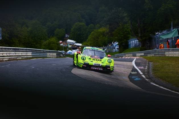 Der neongelbe Porsche 911 GT3 R mit der Startnummer 911 des Teams Manthey durchfährt eine enge Kurve auf der Nordschleife am Nürburgring. Das Fahrzeug Ayhancan Güven & Kevin Estre & Thomas Preining (Manthey) trägt die markanten Sponsoren KÜS, Motul, Red Bull, Veltins und H&R und ist mit einem Gran Turismo-Schriftzug versehen. Im Hintergrund sind Streckenposten in orangefarbener Kleidung sowie Zuschauer am Streckenrand zu sehen. Manthey Porsche 911 GT3 911 auf der Nordschleife am Nürburgring