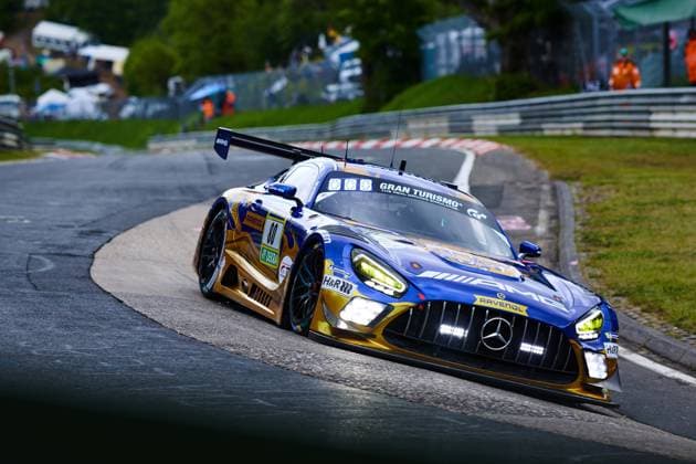 Der Mercedes-AMG GT3 mit der Startnummer 80 des Mercedes-AMG Team RAVENOL durchfährt eine enge Kurve auf der Nordschleife des Nürburgrings. Das Fahrzeug im charakteristischen Blau-Gold-Design mit RAVENOL- und Gran-Turismo-Sponsoring liegt tief in der Kurve, die Scheinwerfer leuchten hell. Sponsoren wie H&R und DEKRA sind deutlich auf der Karosserie erkennbar. Fabian Schiller & Luca Stolz & Maro Engel & Maxime Martin (Mercedes-AMG Team RAVENOL) Mercedes-AMG GT3 80 von RAVENOL in der Kurve am Nürburgring