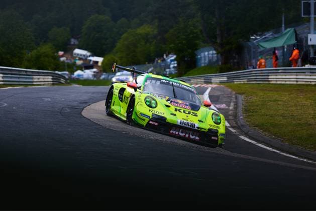 Der auffällige neongelbe Porsche 911 GT3 R mit der Startnummer 911 von Manthey durchfährt eine enge Kurve auf der Nordschleife des Nürburgrings. Das Fahrzeug Ayhancan Güven & Kevin Estre & Thomas Preining (Manthey) trägt prominent die Sponsoren KÜS, Veltins, Motul, Red Bull und H&R und besticht durch sein markantes neongelb-schwarzes Farbdesign. Im Hintergrund sind Streckenposten sowie Zuschauerbereiche zu erkennen. Manthey Porsche 911 GT3 R 911 nimmt Kurve auf der Nordschleife