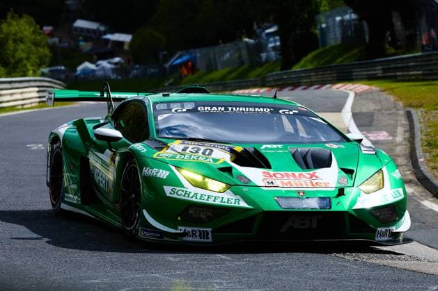 Der grüne Lamborghini Huracán GT3 mit der Startnummer 130 des Red Bull Team ABT durchfährt mit eingeschalteten Scheinwerfern eine Kurve auf der Nordschleife des Nürburgrings. Das Fahrzeug Marco Mapelli & Nicholas Yelloly & Nicky Catsburg (Red Bull Team ABT) trägt die Sponsoren Schaeffler, SONAX Müller und H&R und ist mit Michelin-Reifen ausgestattet. Im Hintergrund sind die charakteristischen Streckenbegrenzungen und das grüne Umfeld der Eifel zu erkennen. Lamborghini Huracán GT3 130 von Red Bull Team ABT auf der Nordschleife