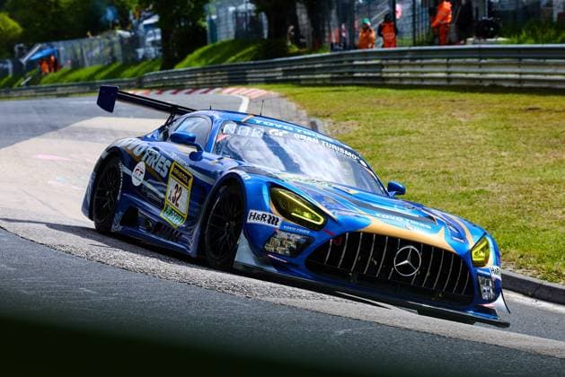 Der Mercedes-AMG GT3 mit der Startnummer 32 des Teams Toyo Tires with Ring Racing durchfährt eine Kurve auf dem Nürburgring. Das Fahrzeug Andreas Gülden & Tim Sandtler & Yuichi Nakayama (Toyo Tires with Ring Racing) präsentiert sich in einem auffälligen blau-goldenen Farbdesign mit Sponsoren wie Toyo Tires, RAVENOL, H&R und Gran Turismo. Im Hintergrund sind Streckenposten in orangefarbener Arbeitskleidung zu erkennen. Mercedes-AMG GT3 32 von Toyo Tires with Ring Racing auf dem Nürburgring