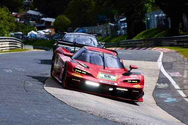 Der rote Lamborghini Huracán GT3 mit der Startnummer 66 von Reiter Engineering durchfährt eine enge Kurve auf der Nordschleife des Nürburgrings. Das Fahrzeug Arne Hoffmeister & Julien Apotheloz & Laurents Hörr & Marcel Marchewicz & Miklas Born (Reiter Engineering) trägt die Sponsoren RAVENOL, DEKRA, H&R und Gran Turismo und ist mit eingeschaltetem Tagfahrlicht unterwegs. Im Hintergrund ist ein weiteres Fahrzeug zu erkennen, während Zuschauer und Hospitality-Zelte die Kulisse der Eifel-Rennstrecke beleben. Lamborghini Huracán GT3 66 von Reiter Engineering auf der Nordschleife