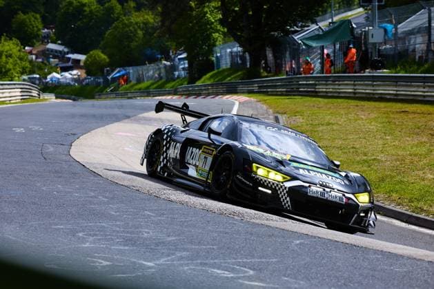 Der Audi R8 LMS GT3 mit der Startnummer 71 von JUTA Racing durchfährt eine enge Kurve auf der Nordschleife des Nürburgrings. Das schwarz-weiß karierte Fahrzeug mit den Sponsoren Alphazoo und RAVENOL ist mit leuchtend gelben Scheinwerfern unterwegs. Im Hintergrund sind Streckenposten in orangefarbener Kleidung sowie Zuschauerbereiche zu erkennen. Björn Großmann & Christer Jöns & Otto Blank & Pierre Kaffer (JUTA Racing) Audi R8 LMS GT3 71 von JUTA Racing auf der Nordschleife