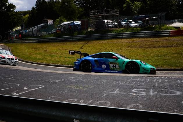 Der Porsche 911 GT3 R (Klaus Bachler & Morris Schuring & Sven Müller & Tim Heinemann (Falken Motorsports)) von Falken Motorsports durchfährt eine Kurve auf der Nordschleife des Nürburgrings. Das Fahrzeug trägt das charakteristische blau-türkise Falken-Design mit den Sponsoren RAVENOL, DEKRA und Falkentyre.com. Im Hintergrund ist ein weiteres Fahrzeug mit H&R-Sponsoring zu sehen. Falken Motorsports Porsche 44 auf der Nordschleife