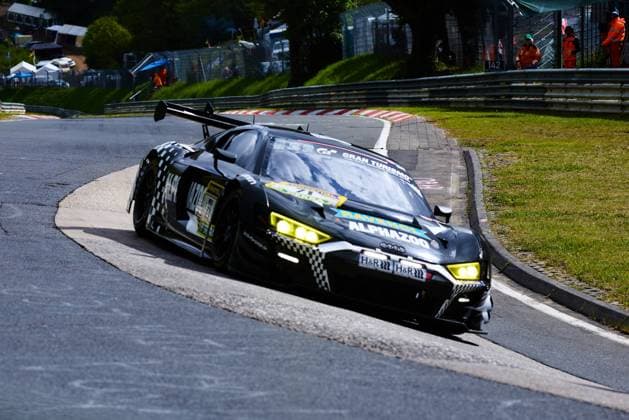 Der schwarze Audi R8 GT3 mit der Startnummer 444 von Adrenalin Motorsport Team Mainhattan Wheels durchfährt eine enge Kurve auf der Nordschleife beim ADAC RAVENOL 24h Nürburgring 2026. Das Fahrzeug im markanten Schachbrettmuster-Design trägt die Sponsoren Alphazoo, RAVENOL, H&R sowie Gran Turismo und nimmt die Kurve mit eingeschalteten Scheinwerfern. Im Hintergrund sind Streckenposten in orangefarbener Sicherheitskleidung sowie der Kilometerstein 142 zu erkennen. 444; Frauenfußball Nationalmannschaft Polen Audi R8 GT3 444 von Adrenalin Motorsport in der Nordschleife-Kurve