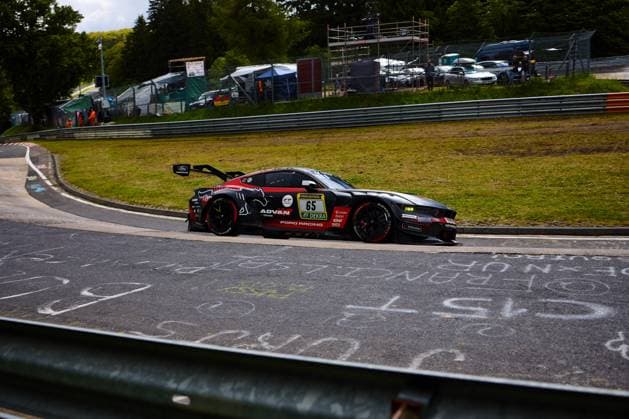 Der Ford Mustang GT3 Colin Caresani & David Schumacher & Hubert Haupt & Vincent Kolb (HRT Ford Racing) von HRT Ford Racing durchfährt eine Kurve auf dem Nürburgring. Das schwarz-rote Fahrzeug mit der Startnummer 65 trägt die Sponsoren ADVAN, RAVENOL und DEKRA und ist mit Ford-Racing-Logos versehen. Im Hintergrund sind Tribünen, Absperrungen sowie Zuschauer zu erkennen, die die ikonische Atmosphäre des 24-Stunden-Rennens unterstreichen. Ford Mustang GT3 65 von HRT Ford Racing in der Kurve am Nürburgring