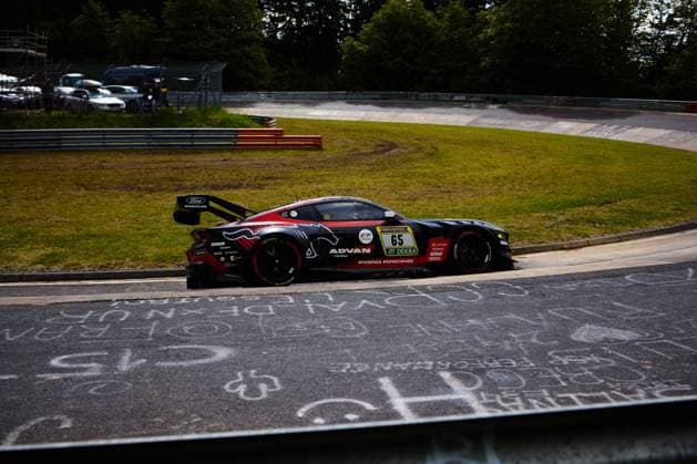 Der Ford Mustang GT3 mit der Startnummer 65 des Teams HRT Ford Racing durchfährt eine Kurve auf dem Nürburgring. Das in Schwarz und Rot gehaltene Fahrzeug trägt prominente Sponsoren-Logos von ADVAN Yokohama und Ford Racing auf der Flanke. Im Hintergrund sind die typischen orangen Curbs und die bewaldete Landschaft der Eifel zu sehen. Colin Caresani & David Schumacher & Hubert Haupt & Vincent Kolb (HRT Ford Racing) Ford Mustang GT3 65 von HRT Ford Racing auf dem Nürburgring