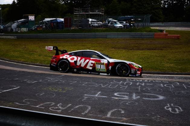 Der BMW M4 GT3 mit der Startnummer 1 von ROWE RACING durchfährt eine Kurve auf dem Nürburgring. Das Fahrzeug trägt das markante rot-weiße ROWE-Design mit RAVENOL- und DEKRA-Sponsoring und ist mit BMW M Motorsport-Branding versehen. Im Vordergrund sind auf dem Asphalt aufgemalte Fangrüße zu sehen, im Hintergrund Fangzäune und Absperrungen der Rennstrecke. Augusto Farfus & Jordan Pepper & Kelvin van der Linde & Raffaele Marciello (ROWE RACING) BMW M4 GT3 1 von ROWE RACING auf dem Nürburgring