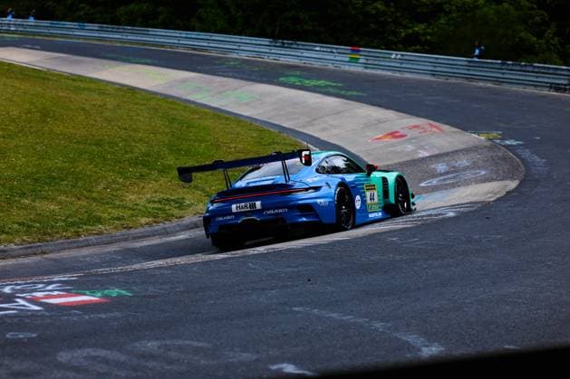 Der Porsche 911 GT3 mit der Startnummer 44 von Falken Motorsports durchfährt eine Kurve auf der Nordschleife am Nürburgring. Das Fahrzeug Klaus Bachler & Morris Schuring & Sven Müller & Tim Heinemann (Falken Motorsports) trägt die charakteristische blau-grüne Falken-Lackierung mit den Sponsoren H&R und RAVENOL und ist mit einem markanten Heckflügel ausgestattet. Die typischen bunten Kreidebilder auf dem Asphalt der Nordschleife sind im Vordergrund zu sehen. Falken Motorsports Porsche 911 GT3 44 auf der Nordschleife