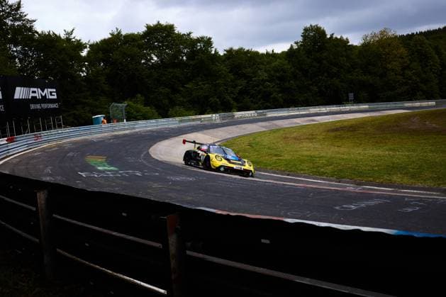 Der gelb-schwarz lackierte Porsche von Reiter Engineering durchfährt eine charakteristische Kurve der Nordschleife am Nürburgring. Im Hintergrund ist das AMG-Banner mit dem Schriftzug Driving Performance zu erkennen. Das Fahrzeug mit der Startnummer 91 nimmt die Kurve auf dem bekannt anspruchsvollen Kurs in der Eifel. Rennfahrzeug Gelb-schwarzer Porsche 91 von Reiter Engineering in der Nordschleife-Kurve