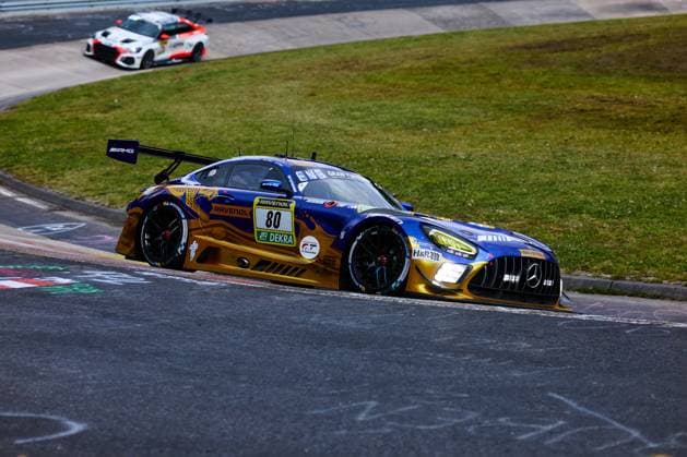 Der Mercedes-AMG GT3 mit der Startnummer 80 des Teams Mercedes-AMG Team RAVENOL durchfährt eine enge Kurve am Nürburgring. Das Fahrzeug Fabian Schiller & Luca Stolz & Maro Engel & Maxime Martin (Mercedes-AMG Team RAVENOL) trägt das charakteristische blau-goldene Farbschema mit den Sponsoren RAVENOL, H&R und DEKRA. Im Hintergrund ist ein weiteres Fahrzeug auf der Strecke zu sehen. Mercedes-AMG GT3 80 von RAVENOL in der Kurve am Nürburgring