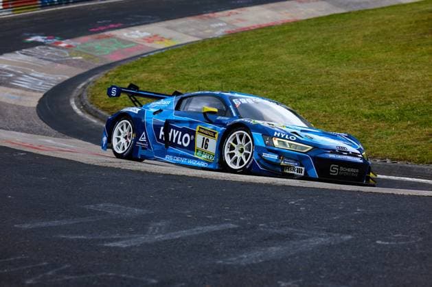 Der Audi R8 LMS GT3 mit der Startnummer 16 von Scherer PHX durchfährt eine Kurve auf dem Nürburgring. Das in leuchtendem Blau gehaltene Fahrzeug Alexander Sims & Ben Green & Christopher Haase (Scherer Sport PHX) trägt die Sponsoren HYLO, RAVENOL und IRS Dent Wizard und ist mit Michelin-Reifen ausgestattet. TÜV Rheinland sowie Scherer Sportarx sind ebenfalls als Partner auf der Karosserie vertreten. Audi R8 LMS GT3 16 von Scherer Sport PHX auf der Nordschleife