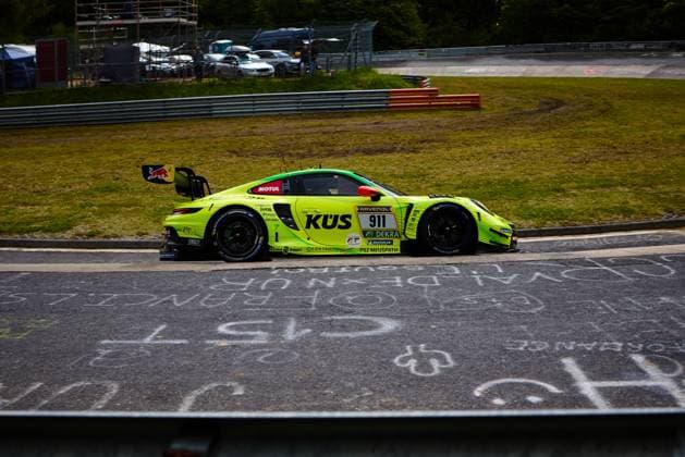 Der leuchtend gelb-grüne Porsche 911 GT3 R mit der Startnummer 911 des Teams Manthey durchfährt die Rennstrecke am Nürburgring. Das auffällig gestaltete Fahrzeug Ayhancan Güven & Kevin Estre & Thomas Preining (Manthey) trägt die Hauptsponsoren KÜS und RAVENOL prominent auf der Karosserie sowie einen Red Bull-Aufkleber am Heckflügel. Im Hintergrund sind Fangzäune, Leitplanken und Fahrzeuge im Fahrerlager erkennbar, während auf dem Asphalt im Vordergrund Fanschriften zu lesen sind. Porsche 911 GT3 R 911 von Manthey im Renneinsatz am Nürburgring