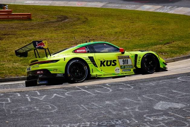 Der neongelbe Porsche 911 GT3 R mit der Startnummer 911 des Teams Manthey durchfährt eine Kurve auf dem Nürburgring. Das auffällig gestaltete Fahrzeug Ayhancan Güven & Kevin Estre & Thomas Preining (Manthey) trägt die Sponsoren KÜS, RAVENOL, Motul, Red Bull und Michelin prominent auf der Karosserie. Der großen Heckflügel mit Red-Bull-Branding und die aerodynamischen Anbauteile unterstreichen den Renncharakter des GT3-Fahrzeugs. Porsche 911 GT3 R 911 von Manthey im Neongelb auf dem Nürburgring
