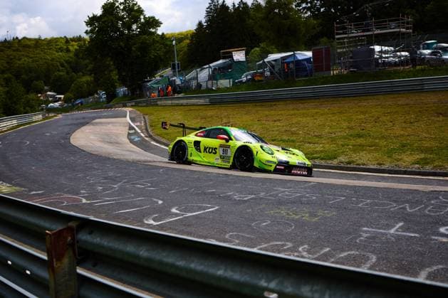 Der auffällig neongelb-grüne Porsche 911 GT3 R mit der Startnummer 911 von Manthey nimmt eine enge Rechtskurve auf der Nordschleife des Nürburgrings. Das Fahrzeug Ayhancan Güven & Kevin Estre & Thomas Preining (Manthey) mit den Sponsoren KÜS, MOTUL und RAVENOL durchfährt die charakteristische Kurve, während im Hintergrund die bewaldeten Eifelhügel und das Fahrerlager sichtbar sind. Porsche 911 GT3 R 911 von Manthey auf der Nordschleife
