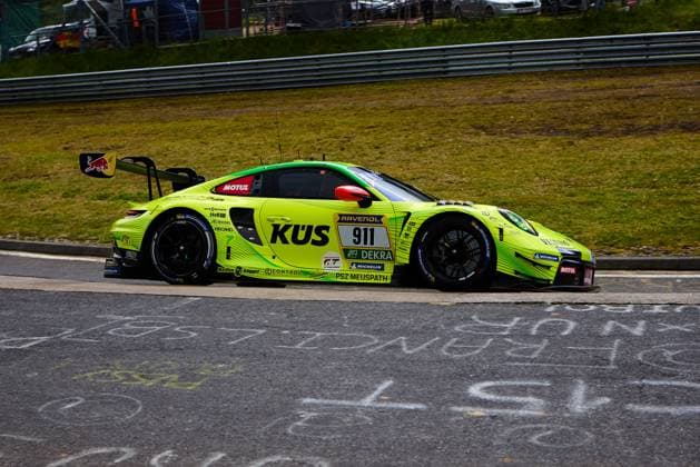 Der Porsche 911 GT3 R mit der Startnummer 911 des Teams Manthey durchfährt eine Kurve am Nürburgring. Das Fahrzeug Ayhancan Güven & Kevin Estre & Thomas Preining (Manthey) präsentiert sich in auffälligem Neongelb-Grün mit dem KÜS-Hauptsponsoring sowie den Logos von RAVENOL, DEKRA, Motul und Red Bull. Auf dem Asphalt sind Fanschriftzüge mit Kreide zu erkennen, die die Atmosphäre des 24-Stunden-Rennens unterstreichen. Porsche 911 GT3 R 911 von Manthey im KÜS-Design am Nürburgring