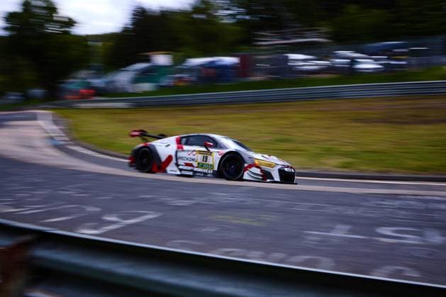 Ein weißer Audi R8 LMS mit der Startnummer 19 durchfährt dynamisch eine Kurve am Nürburgring. Das Fahrzeug trägt ein markantes rot-weißes Design mit den Sponsoren PECKIONI und MOODY S WORKWEAR sowie dem DEKRA-Logo. Die Panning-Aufnahme unterstreicht die hohe Geschwindigkeit des GT-Fahrzeugs beim 24h-Rennen auf der Nordschleife. Audi R8 LMS 19 mit Panning-Aufnahme am Nürburgring
