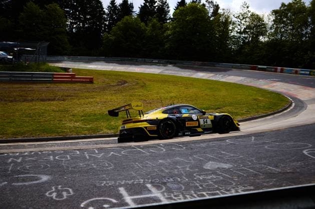 Der Bastian Buss & Michael Christensen & Michael Klitgaard Christensen (Dinamic GT) von Dinamic GT durchfährt eine enge Rechtskurve auf dem Nürburgring. Das in Gelb und Schwarz gehaltene Fahrzeug trägt die Startnummer 54 und ist mit Sponsoren wie Pirelli, RAVENOL und DEKRA versehen. Im Hintergrund ist die typische Nürburgring-Kulisse mit Leitplanken, Curbs und Bäumen sichtbar. Porsche 911 GT3 54 von Dinamic GT in der Kurve am Nürburgring