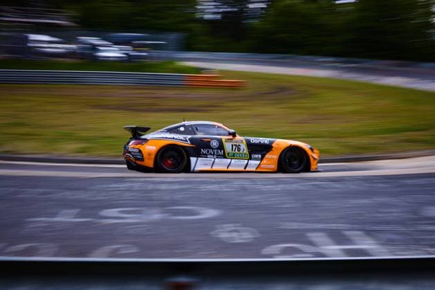 Der Mercedes-AMG GT3 mit der Startnummer 176 von PROsport Racing durchfährt mit hoher Geschwindigkeit eine Kurve auf dem Nürburgring. Das auffällige orange-schwarz-weiße Fahrzeug Abée Anton & Guilherme de Oliveira & Jörg Viebahn & LLuc Ibanez & Yannik Himmels (PROsport Racing) trägt die Sponsoren NOVEM Gold, RAVENOL, Dorint und DEKRA auf dem Livery. Die Panning-Aufnahme betont die Dynamik und Geschwindigkeit des Fahrzeugs während des ADAC RAVENOL 24h Nürburgring 2026. Mercedes-AMG GT3 176 von PROsport Racing auf dem Nürburgring