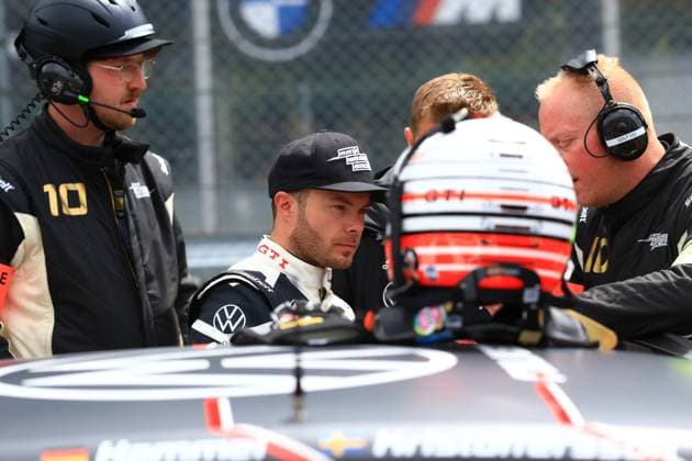 In der Startaufstellung am Nürburgring bereitet sich das Max Kruse Racing Team auf den Rennstart vor. Ein Fahrer im VW GTI-Rennoverall sitzt im Fahrzeug, während Teammitglieder mit Headsets konzentriert letzte Abstimmungen vornehmen. Der Helm eines weiteren Fahrers ist im Vordergrund zu sehen. Am linken Bildrand ist die Nummer 10 auf einem Teamoverall erkennbar, was auf das Max Kruse Racing Fahrzeug 10 hindeutet. Benjamin Leuchter ist ebenfalls im Bild erkennbar. Max Kruse Racing VW GTI am Nürburgring – Fahrerwechsel in der Startaufstellung