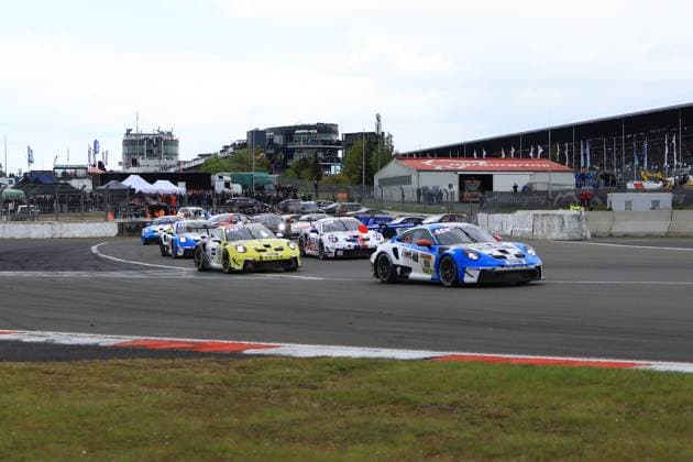 Im Vordergrund führt der blau-weiße Porsche 911 GT3 mit der Startnummer 918 von Mühlner Motorsport powered by H&R das Feld durch eine enge Kurve auf dem Nürburgring-Grand-Prix-Kurs. Dicht dahinter drängt der gelb-schwarze Porsche 911 GT3 mit der Nummer 992, gefolgt von weiteren GT-Fahrzeugen in einer dichten Gruppe. Im Hintergrund sind die Haupttribüne sowie das AMG-Logo auf dem Kontrollturm des Nürburgrings zu erkennen. Das Bild zeigt die intensive Anfangsphase des ADAC RAVENOL 24h Nürburgring 2026 bei bewölktem Wetter. Startphase beim 24h Nürburgring – Porsche 918 führt das Feld an