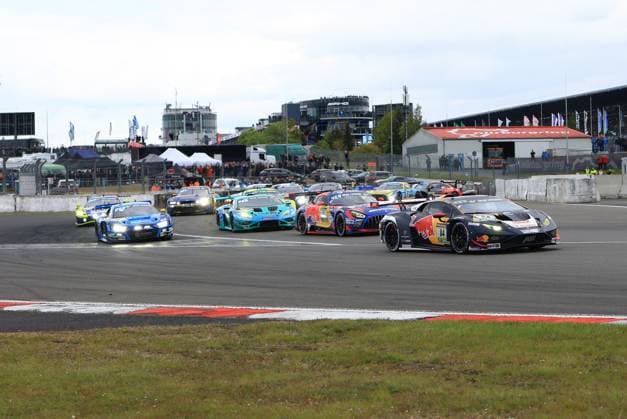 Der Lamborghini Huracán GT3 mit der Startnummer 84 von Red Bull Team ABT führt ein dichtes Starterfeld in einer Kurve des Nürburgrings an. Im Hintergrund sind zahlreiche weitere GT3-Fahrzeuge im Kampf um Positionen zu sehen, darunter ein blauer Audi, ein türkiser Lamborghini sowie ein Mercedes-AMG GT3. Zuschauer säumen die Strecke und im Hintergrund ist die Nürburgring-Infrastruktur mit dem AMG-Hospitality-Bereich deutlich erkennbar. Luca Engstler & Mirko Bortolotti & Patric Niederhauser (Red Bull Team ABT) Lamborghini 84 Red Bull ABT führt das Starterfeld am Nürburgring