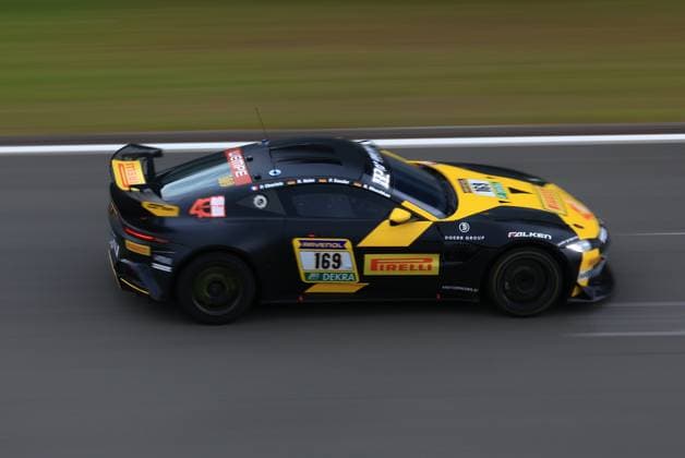 Der Heiko Hahn & Philippe Charlaix & Roland Waschkau & Peter Sander (Doerr Motorsport) von Doerr Motorsport rast mit hohem Tempo über den Nürburgring. Das schwarz-gelbe Fahrzeug, ein Aston Martin GT4, trägt die Startnummer 169 und ist mit Sponsoren wie Pirelli, Falken, RAVENOL und DEKRA dekoriert. Die Panning-Aufnahme betont die Dynamik des Rennwagens auf der Strecke. Aston Martin 169 von Doerr Motorsport auf dem Nürburgring