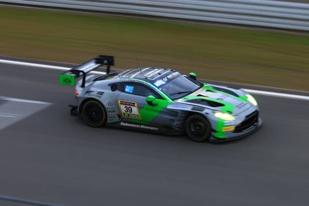 Der Henry Walkenhorst & Anders Buchardt & Mex Jansen & Nico Hantke (Walkenhorst Motorsport) von Walkenhorst Motorsport ist mit hoher Geschwindigkeit auf der Rennstrecke des Nürburgrings unterwegs. Das Fahrzeug in der charakteristischen grau-grünen Lackierung mit leuchtend grünen Akzenten zeigt Bewegungsunschärfe, die die Dynamik des Rennens eindrucksvoll unterstreicht. Die eingeschalteten Scheinwerfer deuten auf Dämmerungs- oder Nachtbedingungen hin. Sponsoren wie RAVENOL und DEKRA sind deutlich auf der Fahrzeugseite zu erkennen. Aston Martin Vantage GT3 39 von Walkenhorst Motorsport auf dem Nürburgring