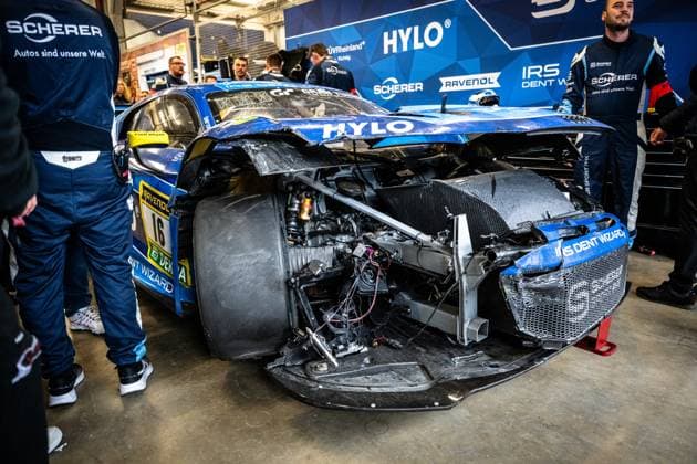 Das Alexander Sims & Ben Green & Christopher Haase (Scherer PHX) von Scherer Sport PHX steht nach einem schweren Unfall in der Boxengarage am Nürburgring. Die Front des blauen GT3-Fahrzeugs ist vollständig zerstört: Karosserieteile fehlen, Kühlerleitungen, Kabel und Fahrwerkskomponenten liegen frei. Der Frontbereich ist so stark deformiert, dass das Fahrzeug kaum noch als rennfähig erscheint. Teammitglieder in blauen Scherer-Overalls stehen rings um das Fahrzeug. Sponsoren wie HYLO, RAVENOL, IRS Dent Wizard, DEKRA und Cam Shaft sind noch am stark beschädigten Fahrzeug erkennbar. Schwer beschädigter GT3 16 von Scherer Sport PHX in der Box Copyright: xMichaelxGelhardtx