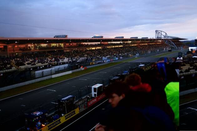 Die Haupttribüne am Nürburgring ist beim ADAC RAVENOL 24h Nürburgring 2026 dicht besetzt. In der Abenddämmerung leuchten die BMW M Power-Werbebanner in der überdachten Tribüne, während Tausende von Zuschauern das Renngeschehen verfolgen. Im Hintergrund ist das charakteristische Achterbahn-Konstrukt des Freizeitparks sowie der bewölkte Abendhimmel über der Eifel zu sehen. Vollbesetzte Haupttribüne am Nürburgring bei Abenddämmerung