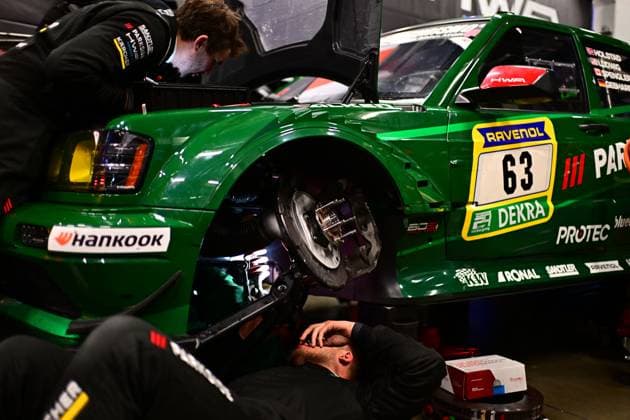 Der grüne Rennwagen Adam Adelson & James Green & Lance David Arnold & Renger Van der Zande (HWA Engineering Speed) von HWA Engineering Speed steht in der Boxengarage am Nürburgring auf Hebeböcken. Zwei Mechaniker arbeiten konzentriert an der freiliegenden Bremsscheibe der Vorderachse – einer kniet darunter, der andere beugt sich von oben herunter. Die Bremsanlage ist vollständig demontiert und gut sichtbar. Sponsoren wie Hankook, RAVENOL, DEKRA und PROTEC sind auf der Karosserie zu erkennen. Auf dem Fahrzeugdach sind die Fahrernames Holstad, Ludwig, Spengler und Gebhardt verzeichnet. HWA Engineering Speed 63 – Mechaniker bei Bremsarbeiten in der Box Copyright: xMichaelxGelhardtx